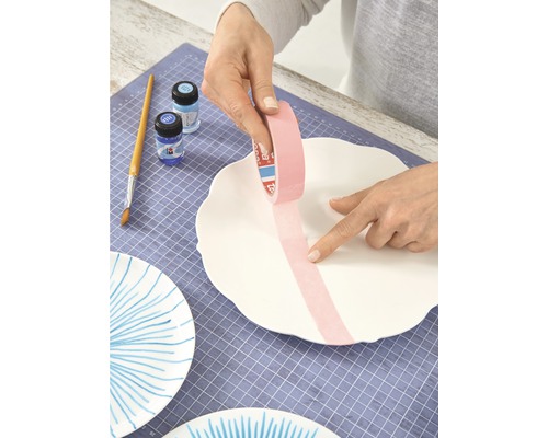 Une personne colle du ruban de masquage sur une assiette en céramique blanche pour la décorer. Pinceau, couleurs et autres assiettes peintes sont visibles.