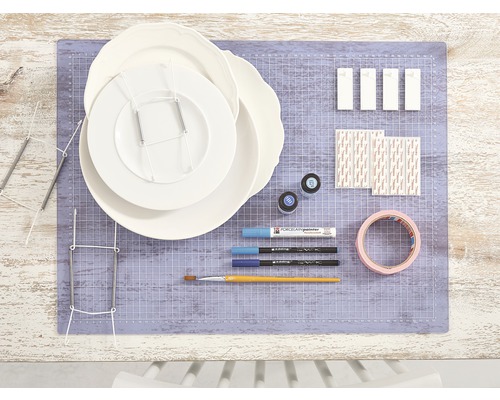 Présentation avec assiettes, supports d''assiettes, peinture pour porcelaine, pinceaux, ruban adhésif et matériel de fixation sur un tapis de découpe
