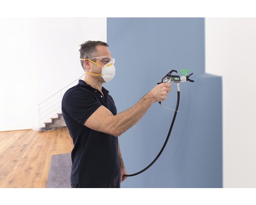 Mann lackiert Wand mit Farbsprühgerät, er trägt Schutzmaske und Schutzbrille