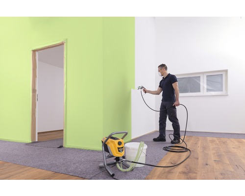 Mann sprüht Farbe mit einer Farbspritzpistole in einem Zimmer. Im Vordergrund steht ein Farbspritzgerät.