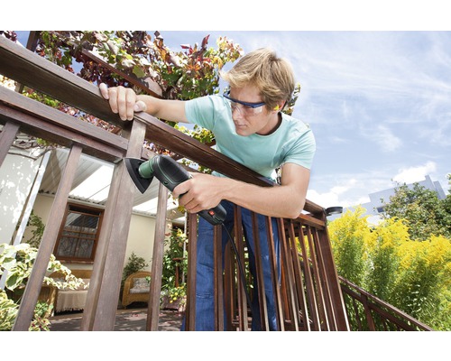 Un homme ponce une balustrade en bois avec une ponceuse multifonction à l''extérieur. Il porte des lunettes de protection.
