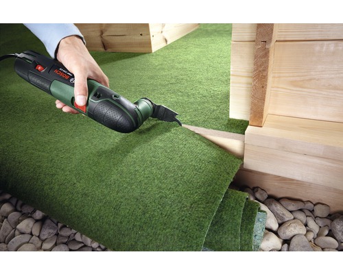 Une personne coupe du gazon synthétique avec un outil multifonction.