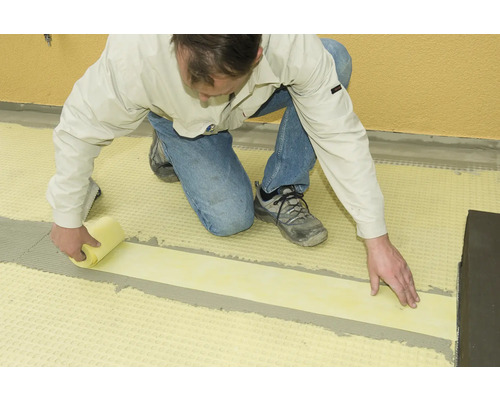 Un homme pose des carreaux avec une bande d''étanchéité jaune et une éponge.