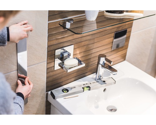 Salle de bain lumineuse avec lavabo, porte-savon, étagère en verre et personne fixant un miroir