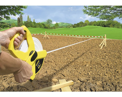 Une personne utilise un ruban à mesurer de 60 mètres de la marque Stanley pour mesurer un terrain délimité par des rubans rouge et blanc.