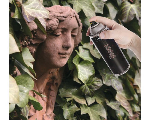 Une main vaporise une statue en pierre avec une bombe de peinture effet rouille devant un fond de lierre.