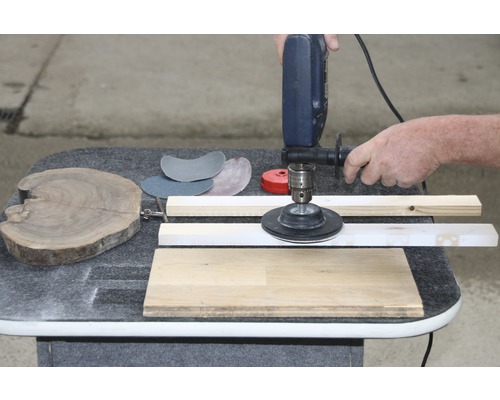 Eine Person verwendet eine Schleifmaschine an einem Holzstück, mit Schleifpapier und Holzlatten auf einem Tisch.