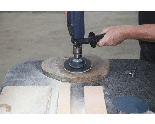 Eine Person schleift ein Holzstück mit einer Schleifmaschine. Schleifpapier und anderes Holz liegen auf dem Tisch.