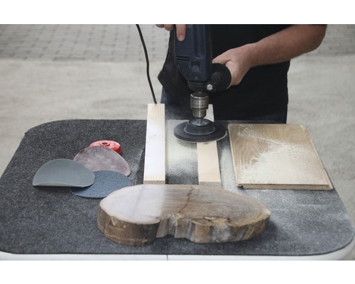 Eine Person schleift Holz mit einem Elektrowerkzeug auf einer Werkbank, Schleifpapier liegt daneben.