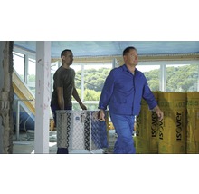 Zwei Handwerker tragen einen Werkzeugkasten und Dämmmaterial in einem Rohbau.