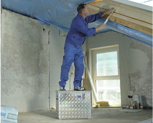 Ein Mann steht auf einer Werkzeugkiste und misst mit einem Zollstock ein Dachgeschoss aus.