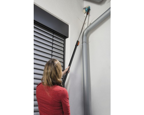 Eine Frau benutzt eine Teleskopbürste zur Reinigung einer Fassade neben einer Regenrinne.