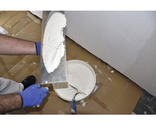 Un artisan applique du plâtre sur un mur avec une spatule. Un seau de plâtre est prêt.