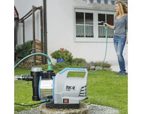 Gartenansicht mit Wasserpumpe und Frau beim Bewässern des Rasens