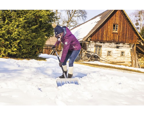Frau schaufelt Schnee mit einer Schneeschaufel vor einem Haus.