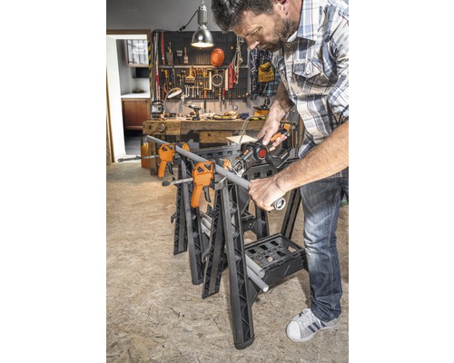 Un homme scie un tuyau sur un établi avec des serre-joints et une scie sabre dans un atelier.