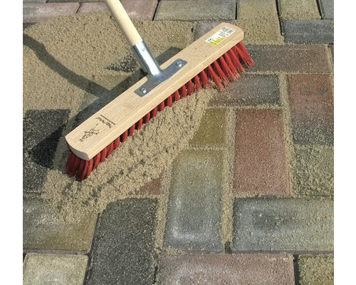 Une brosse de surface à poils rouges balaie le sable dans les joints des pavés.