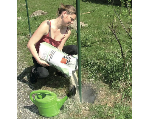 Frau schüttet Beton Schnellzement in ein Loch neben einem Zaunpfosten, mit einer grünen Gießkanne im Vordergrund.