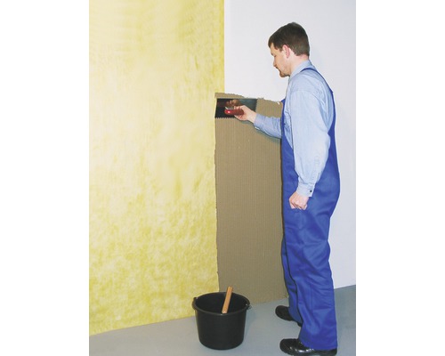 Ein Mann trägt Fliesenkleber mit einer Glättkelle auf eine Wand auf. Ein Eimer und ein Holzstab stehen auf dem Boden.