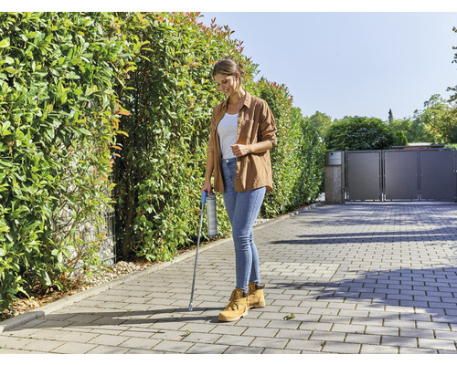 Frau verwendet Unkrautvernichter mit Sprühlanze auf Pflastersteinen vor einer Hecke.