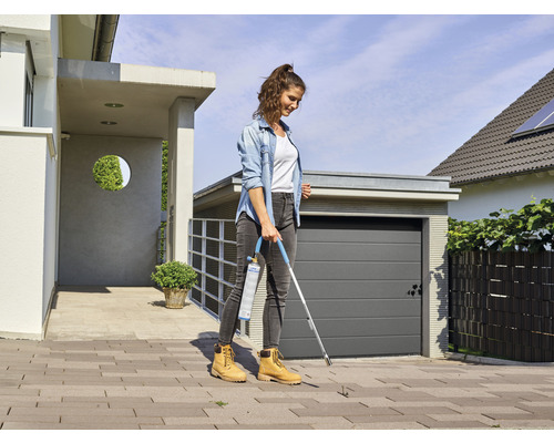 Frau entfernt Unkraut mit einem Gasbrenner auf einer gepflasterten Fläche vor einem Haus.