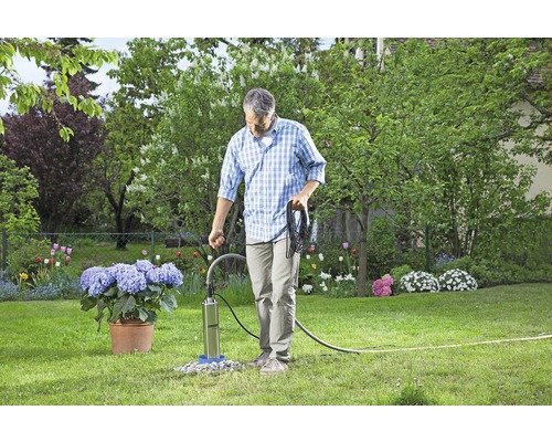 Mann bedient eine Tiefbrunnenpumpe im Garten, umgeben von Blumen und Grünflächen.
