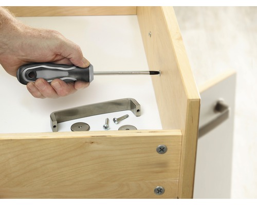 Eine Person befestigt einen Möbelgriff mit einem Schraubendreher an einer Schublade. Montagematerialien liegen bereit.