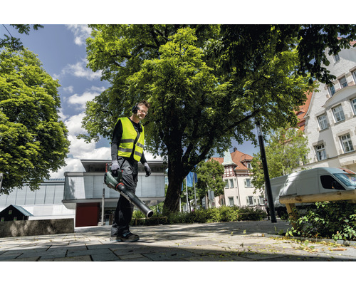 Un homme portant un gilet de sécurité et un casque antibruit utilise un souffleur de feuilles à l''extérieur.