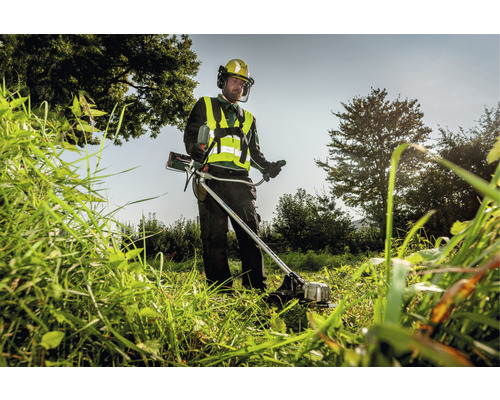 Un homme coupe de l''herbe haute avec une débroussailleuse tout en portant des vêtements de protection.