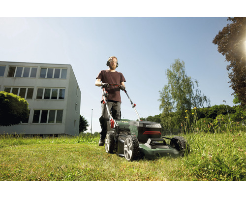 Un homme tond la pelouse devant un bâtiment avec une tondeuse à gazon Metabo.