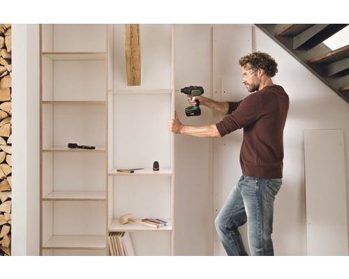 Un homme monte une étagère avec une visseuse sans fil dans une pièce lumineuse.