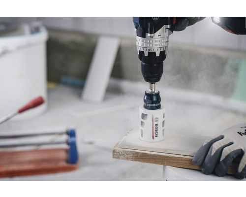 Une perceuse avec un embout de scie cloche perce un trou dans une pièce. Logo Bosch.