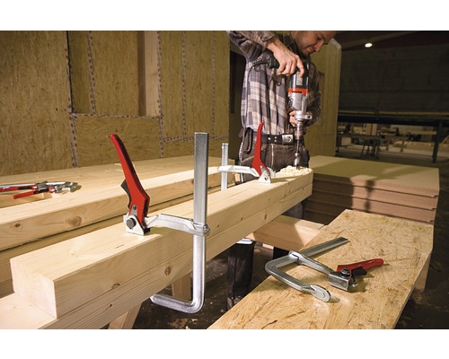 Un homme perce avec une perceuse un morceau de bois fixé avec des serre-joints