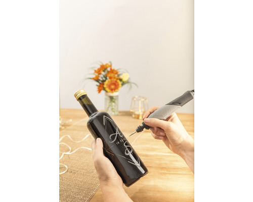 Une personne grave une bouteille en verre avec des initiales et une date à l''aide d''un outil de gravure Dremel.