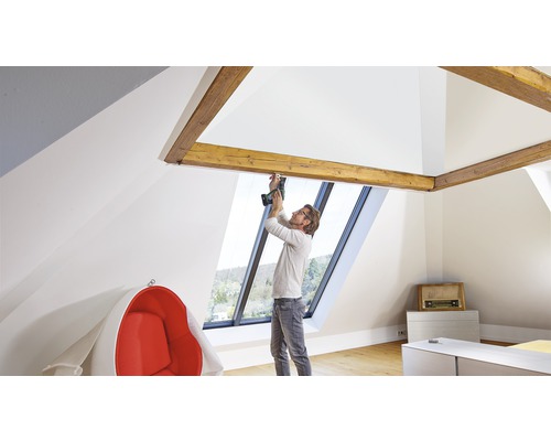 Un homme monte des ferrures sur une fenêtre de toit avec une visseuse sans fil dans une chambre mansardée lumineuse.