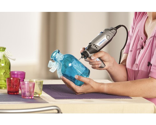 Femme gravant un verre bleu avec un outil multifonction Dremel. Verrerie décorative en arrière-plan.