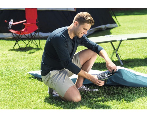 Un homme utilise une pompe à air pour gonfler un matelas pneumatique dans un camping. Une tente et une chaise de camping sont visibles à l''arrière-plan.