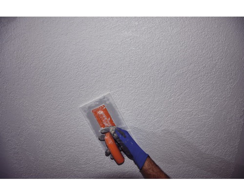 Un homme porte des gants et tient une taloche devant un mur texturé.