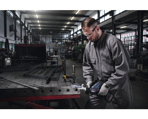 Mann mit Schutzbrille und Gehörschutz arbeitet mit einer Akku-Säbelsäge an einem Metallstück in einer Werkstatt.
