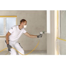 Un homme utilise un pulvérisateur de peinture dans une pièce pour peindre les murs. Il porte des vêtements de protection.