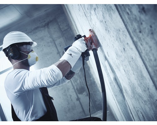 Un ouvrier du bâtiment portant un casque de protection, une protection auditive, des lunettes de protection et un masque respiratoire utilise une rainureuse à béton sur un mur en béton.