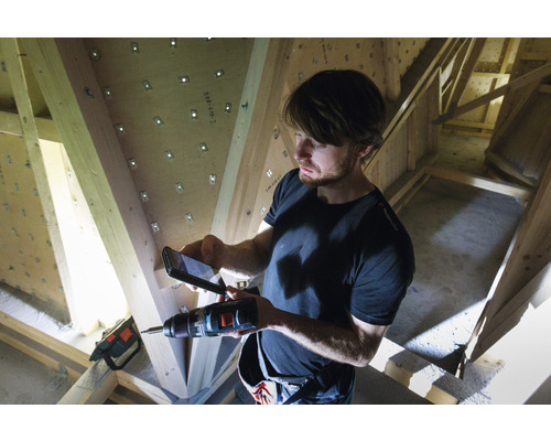 Un homme photographie avec un téléphone portable une perceuse devant une charpente en bois