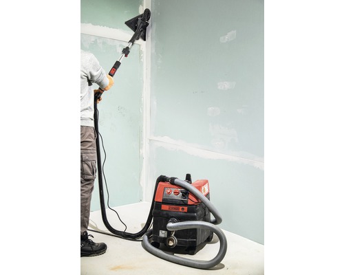 Une personne ponce un mur avec une ponceuse à plaques de plâtre et un aspirateur.
