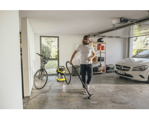 Un homme aspire un garage avec un aspirateur multi-usage, dans lequel se trouvent un vélo, des étagères, une voiture et une porte de garage.