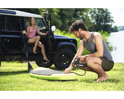 Un homme gonfle un stand-up paddle avec une pompe électrique, avec une femme et un véhicule tout-terrain en arrière-plan.