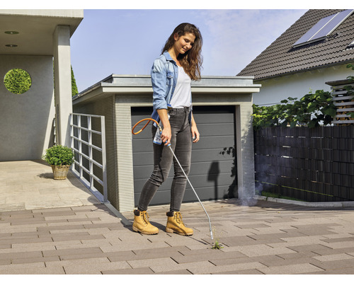 Frau entfernt Unkraut mit einem Gasbrenner auf einer gepflasterten Einfahrt vor einem Haus.