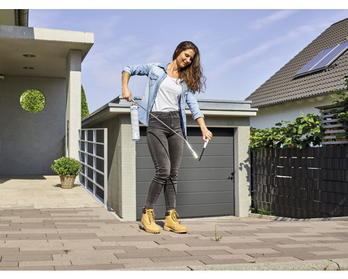 Frau entfernt Unkraut mit einem Gasbrenner auf einer gepflasterten Fläche vor einem Haus.