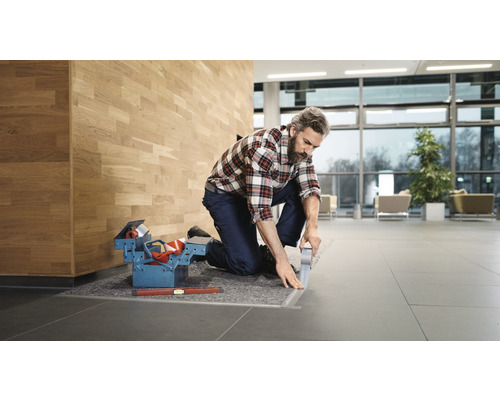 Handwerker kniet auf Fliesenboden und arbeitet an einem Übergangsprofil. Werkzeugkiste und Wasserwaage liegen bereit.