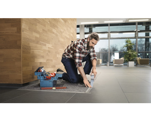 Handwerker kniet und fixiert eine Metallschiene mit Klebeband. Werkzeugkasten und Wasserwaage liegen bereit.