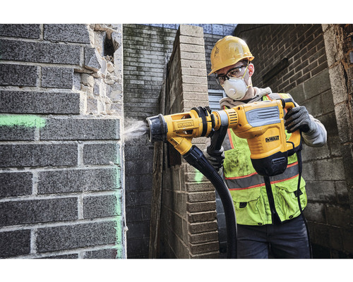 Un ouvrier du bâtiment, équipé de protections, utilise un marteau-piqueur pour travailler sur un mur de briques. L''appareil est équipé d''un dispositif d''aspiration de poussière. Logo DeWalt.
